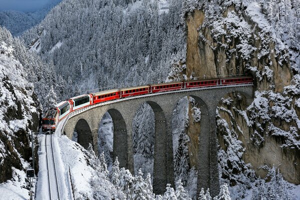 The red train leaves the tunnel in the rock on a narrow high bridge standing among the snow-covered mountain forest