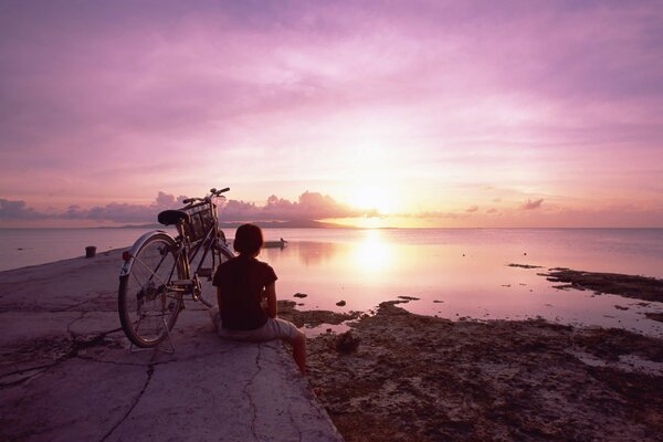 Una niña con una bicicleta en el fondo de una puesta de sol rosa en el reflejo del agua