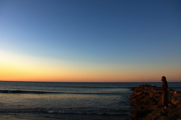 A girl on a rocky seashore watches the sunset