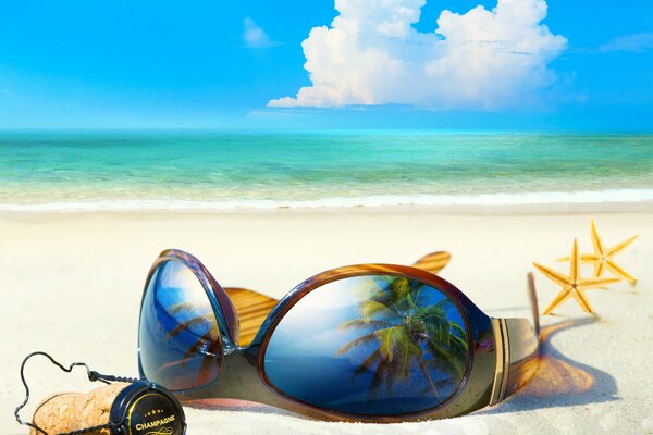 На пляже на белоснежном песке лежат очки солнечные и пробка