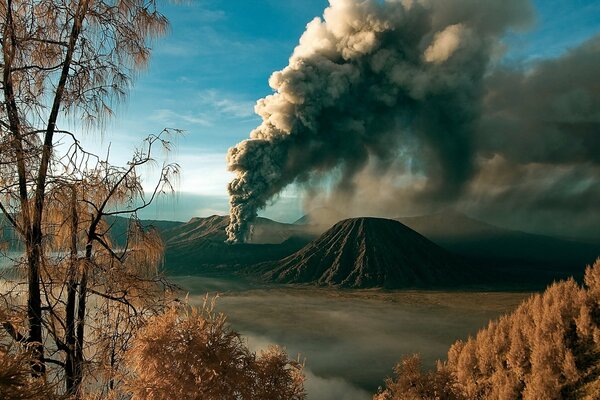 A landscape with a volcano and clouds of smoke against the background of an autumn forest and a bright blue sky