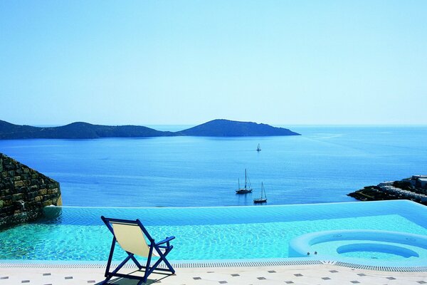 La silla junto a la piscina azul se encuentra cerca del mar, donde los yates y las montañas se ven y el mar se fusiona con el cielo