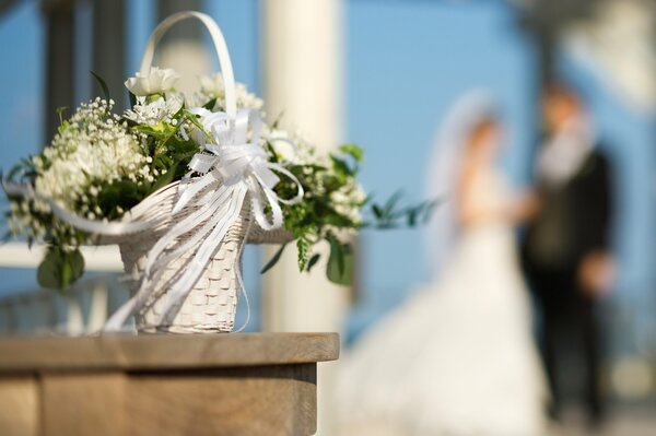 Свадебная белая цветочная корзина с белой лентой на фоне размытых жениха и невесты