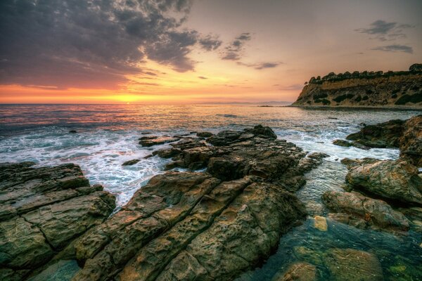 The rocky shore by the foam waves, tinted with a yellow-orange strip of the setting sun covered with brown clouds