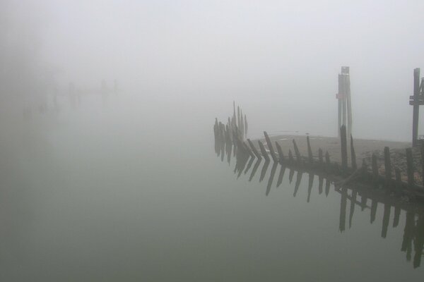 The shore is shrouded in fog. There is a broken fence nearby