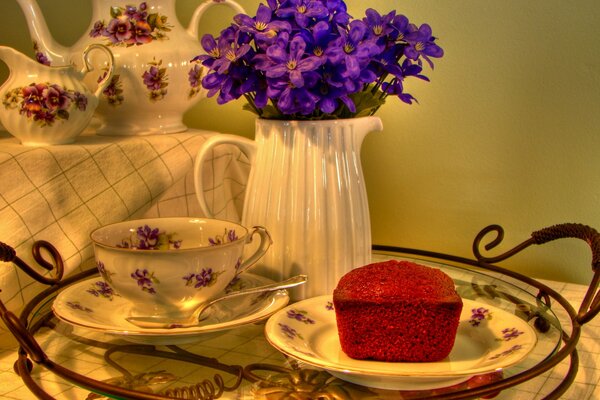 The table has a tablecloth on it there are flowers a tray and a tea set