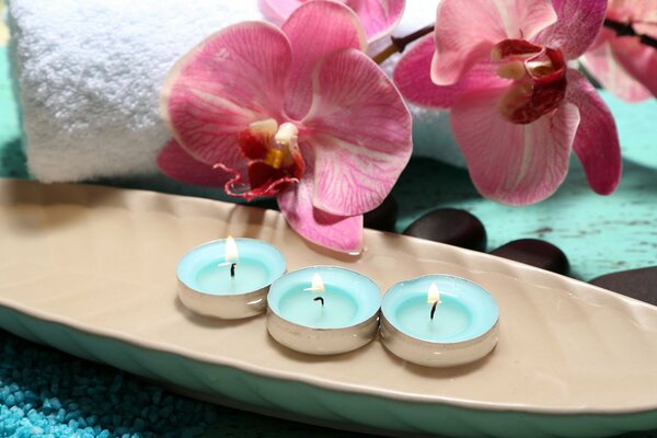 Three burning candles. In the background there is a white towel and orchids