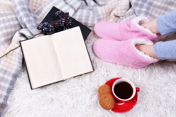 Pies de niña en UGGs rosados caseros. Taza roja con café en un platillo rojo. Galletas de avena. Libro abierto. Manta a cuadros cálida y alfombra blanca con gran siesta