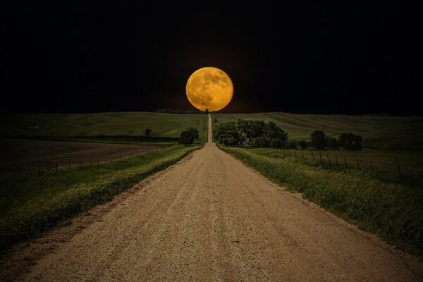 A long road in a field with a round moon turning yellow on the horizon