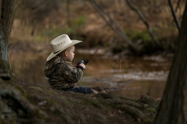 Un niño con un sombrero con una caña de pescar se sienta en la orilla