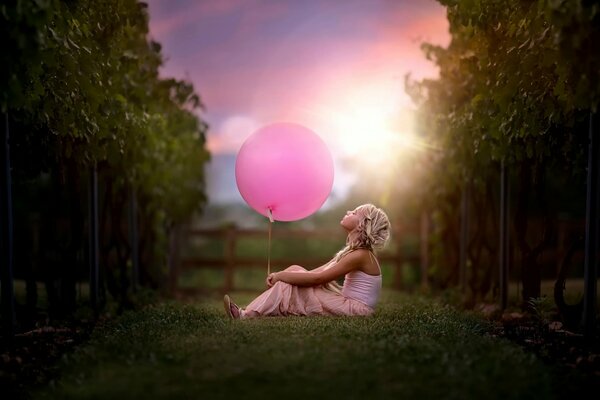 Забавная девочка сидит на траве и держит розовый шарик