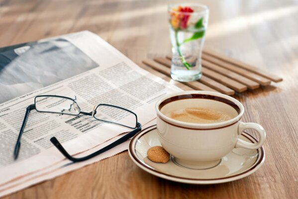 A cup of coffee with a small cookie is on the table, next to it are: a newspaper, glasses and a glass with a flower