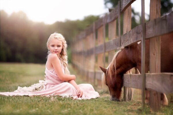 Ein lustiges Mädchen sitzt auf dem Rasen, während ein Pferd hinter einem Zaun grast