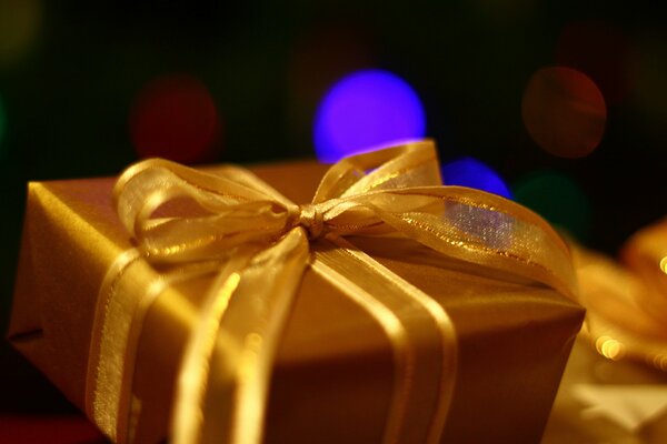Boîte-cadeau dans un emballage doré avec un arc doré sur fond de taches de lumière multicolores