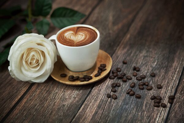 A white rose on the table with a white cup filled with foamy coffee with fragrant coffee beans next to it