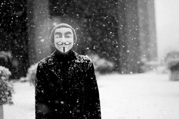 A man in a Guy Fawkes mask under a snowfall