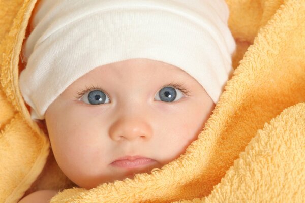 A baby in a white cap is wrapped in a yellow towel