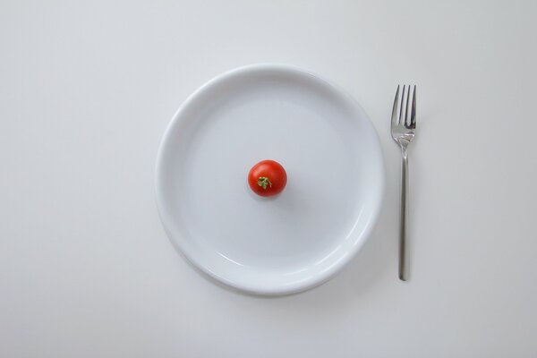 En la mesa blanca, un plato blanco con un tomate Cherry y un tenedor