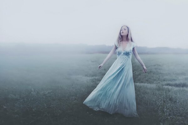 A girl in a long dress stands in the fog on the field