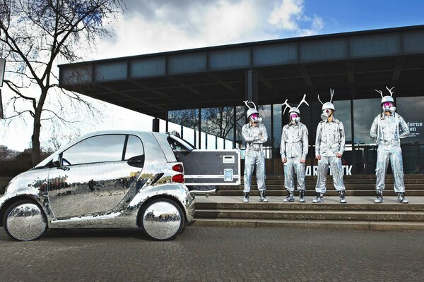 People in shiny overalls near a silver car