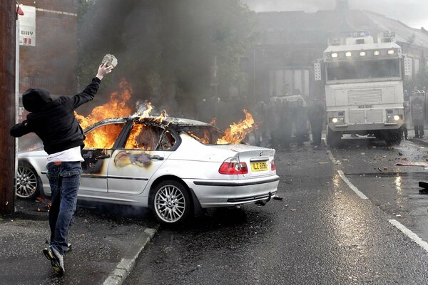 Un matón lanza una piedra a un coche en llamas