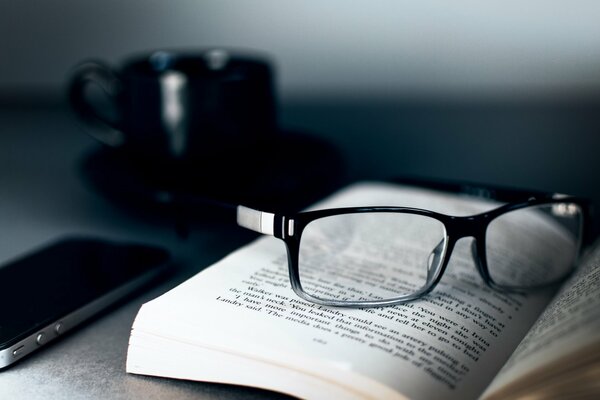 A black cup next to a book, glasses and an iphone 4