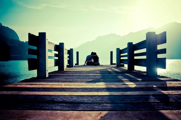 A couple is sitting on a pier against the background of the sea and mountains