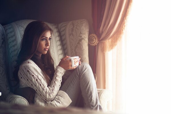 Mädchen in einem Sessel mit einem Becher auf einem Fensterhintergrund