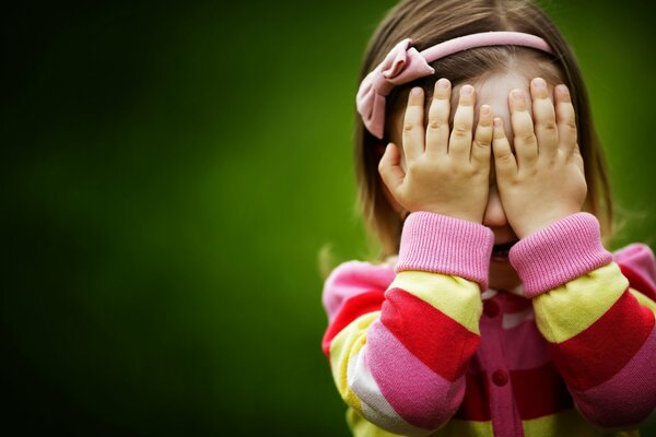 A girl with a pink bow in her hair opens her face with her hands on a green background