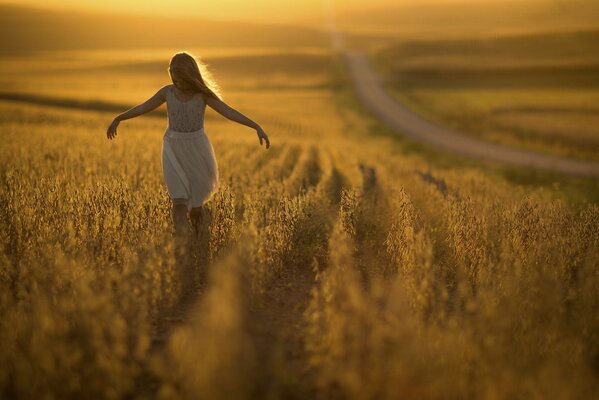 The girl runs with her arms outstretched across the field under the setting sun and in the distance there is a road