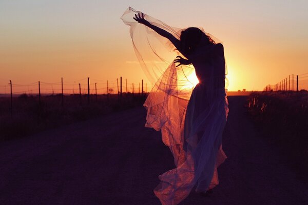 A slender girl in a gauze dress dances on the road against the background of an evening sunset