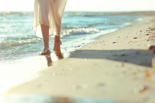 Bare feet of a girl walking on the beach in a white dress