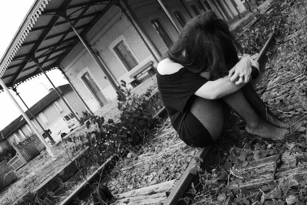 A sad girl in a black dress and tights is sitting on the rails at the old train station waiting for a train