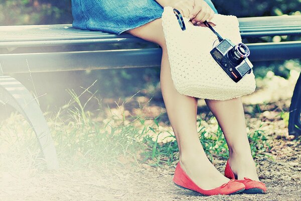 Gambe in ballerine rosse della ragazza su una panchina in gonna di jeans con la macchina fotografica e borsa lavorata a maglia in mani