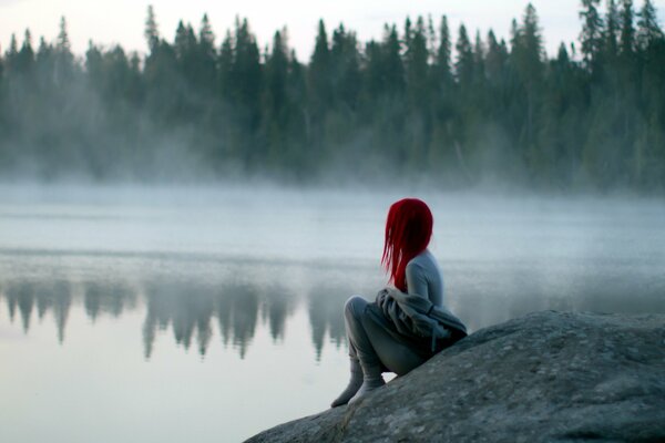 A red-haired girl sits on the shore and looks into the fog