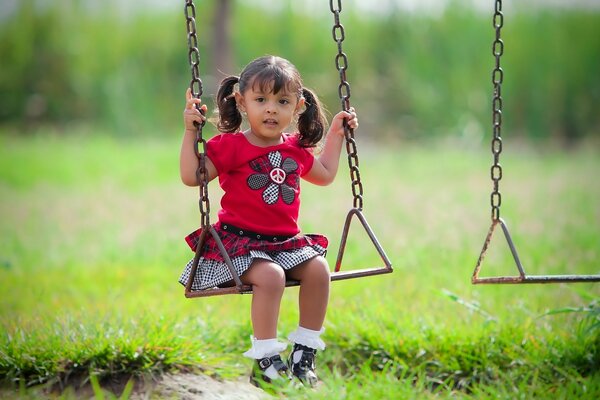 A little brown-haired girl with ponytails in a bright red dress with a flower is sitting on a swing among the greenery on a sunny summer day