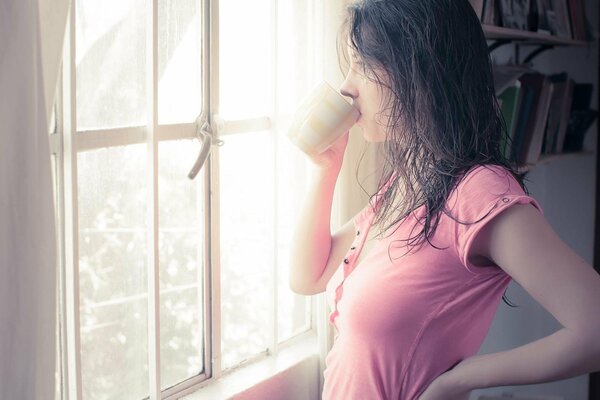 Fille brune avec les cheveux mouillés dans un t-shirt rose boit de la tasse près de la fenêtre