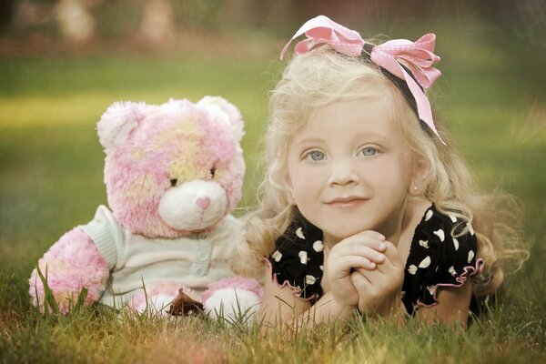 A cute blonde girl with a pink bow and a black dress with white speckles is lying on the grass in the company of a pink and yellow bear cub in a pale blue blouse