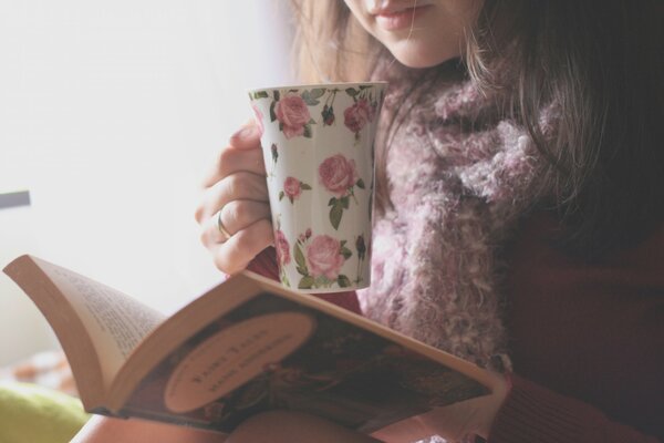 Девушка в свитере читает книгу и пьёт чай