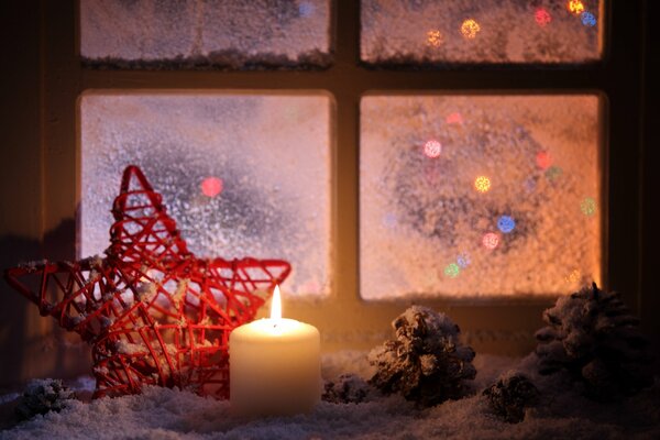 A red braided star and a burning candle with cones on the winter windowsill