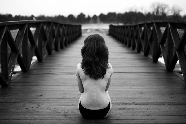 Morena en bragas negras se sienta de espaldas al espectador en un puente de madera y Mira a lo lejos el río y el bosque