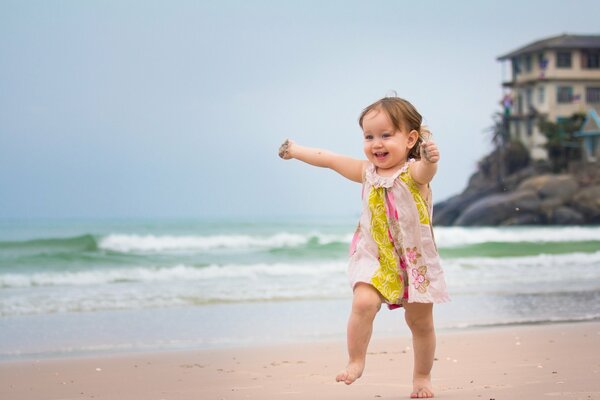 Szczęśliwa dziewczynka w różowo-żółtej sukience z kwiatami kroczy po mokrym wybrzeżu, a jej ręce są poplamione piaskiem