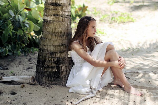 Młoda smutna zamyślona dziewczyna w białej sukience siedzi w cieniu pod palmą wśród nasłonecznionego piasku obejmując kolana