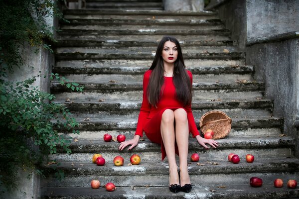Inese Stoner in a red dress, sitting on a staircase strewn with apples