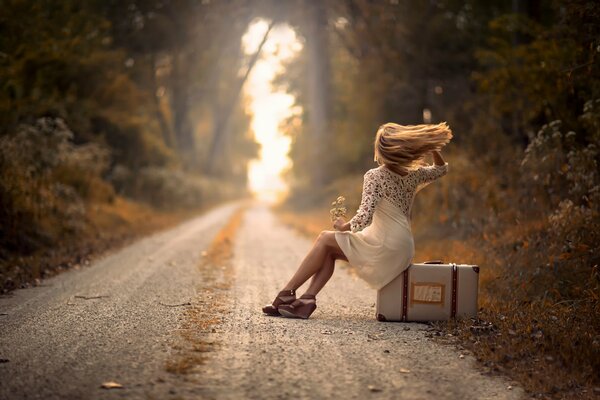 La jeune fille, assise sur une valise sur le bord de la route. Le vent ondule ses cheveux