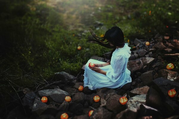 Девушка в белом платье сидящая на камнях с яблоком в руке