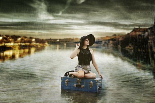 A girl in a hat is sitting on a suitcase in the water