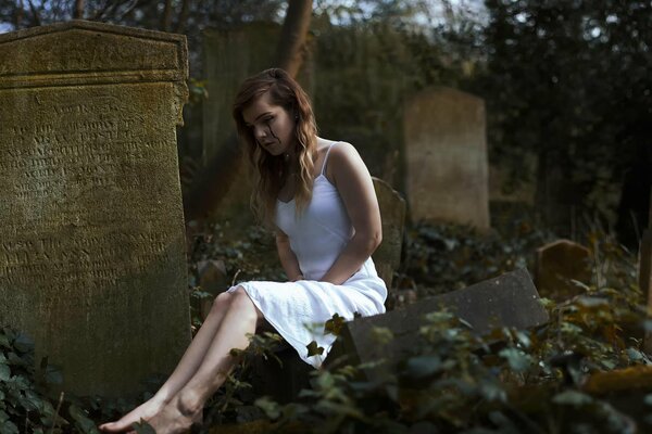 A crying girl in a white dress in the cemetery at the tombstone