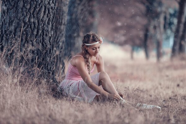 A ballerina girl sits by a tree, puts on pointe shoes