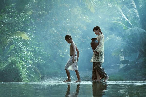 A girl with a jug and a boy walk along a river in the jungle